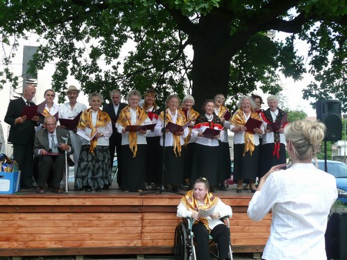 Chór seniorów śpiewa na scenie plenerowej pod drzewem, prowadzony przez dyrygentkę