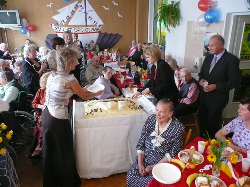 Uroczystość XV lat DPS Oława, seniorzy przy stołach, na środku krojony jest duży tort"